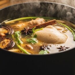 Close-up of clear, golden-hued chicken pho broth simmering gently in a large stockpot with spices and charred aromatics floating, showing the slow, deep flavor extraction process.