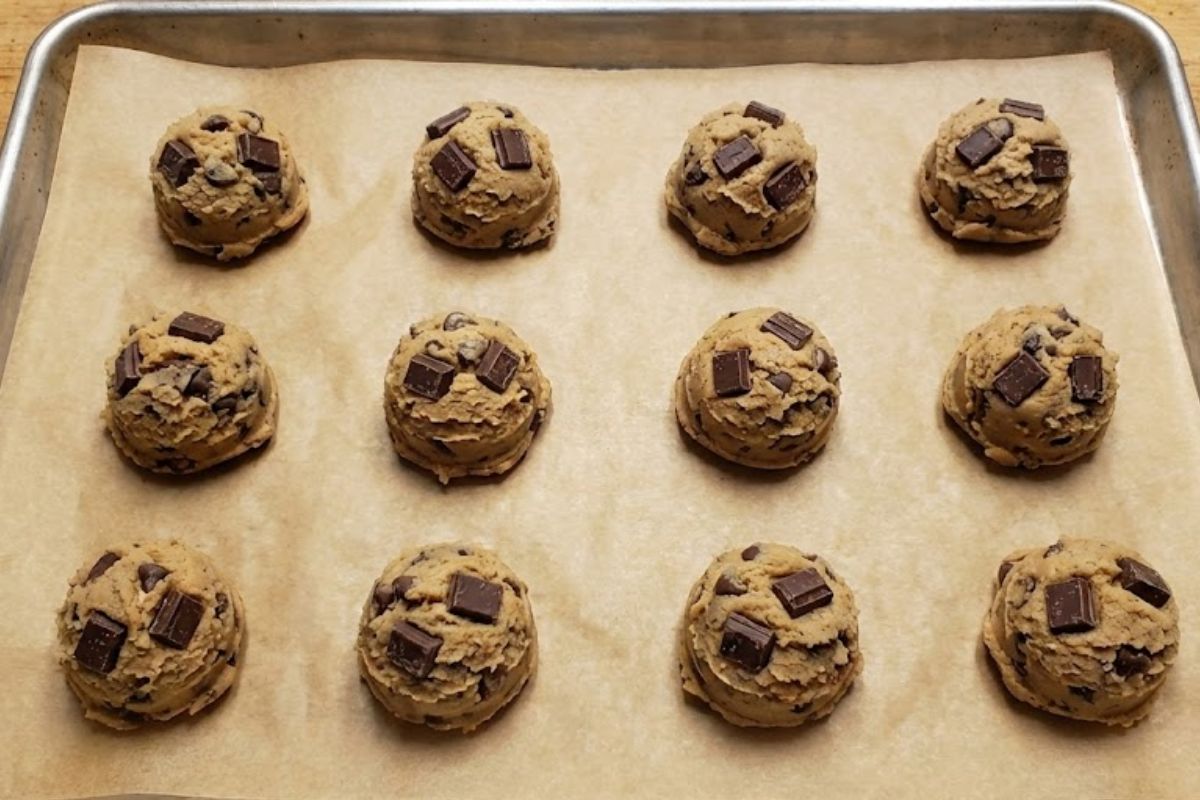 Top-down view of raw chocolate chip cookie dough scoops containing large chocolate chunks arranged on a parchment-lined baking sheet.