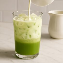 Pouring milk into matcha to create layered pistachio matcha latte in a clear glass