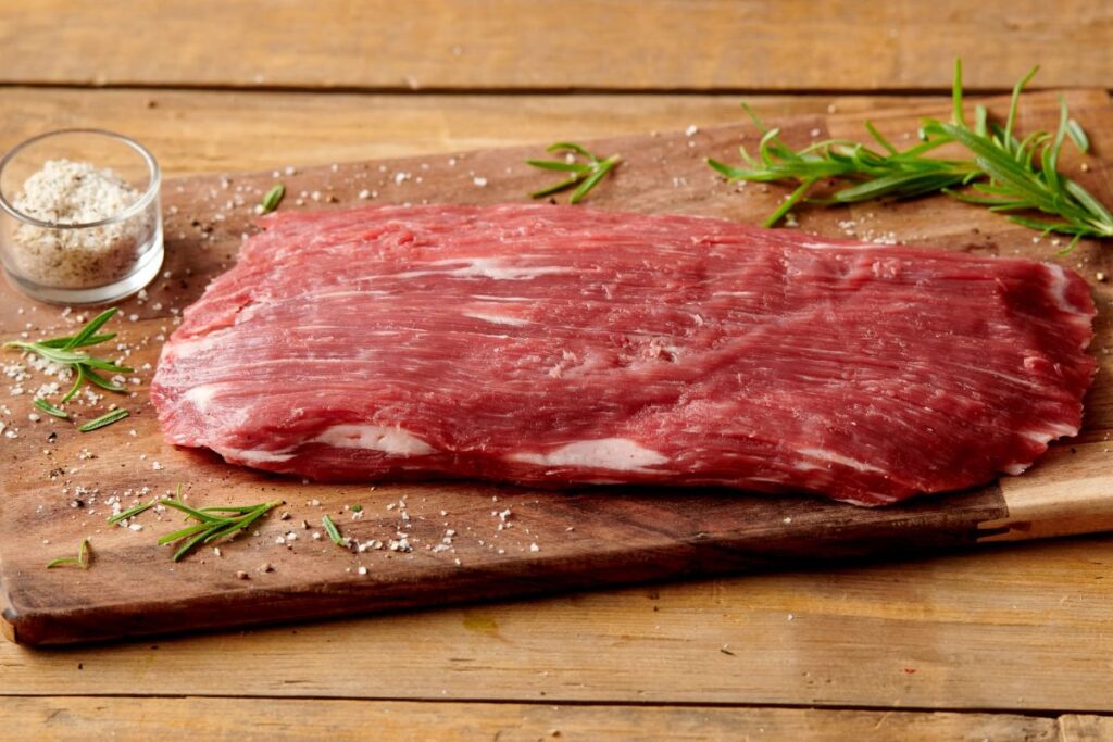 A raw flank steak resting on a wooden cutting board, showcasing the long muscle fibers for slicing against the grain for 