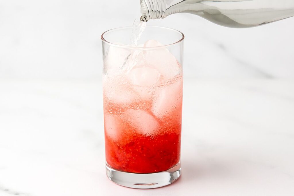 Side-view of cold water being poured slowly into a tall glass over ice and a red strawberry base, creating a beautiful gradient effect.