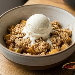 A single serving of the finished, baked Chai-Spiced Apple Crumble (from image_4.png) presented in a rustic handmade ceramic bowl. A scoop of vanilla bean ice cream is melting quickly over the bubbling, golden-brown oat-and-butter topping, highlighting the complex textures of this best fall dessert.