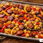 Roasted smoked sausage slices with charred bell peppers and golden potatoes on a baking sheet.